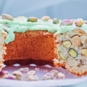 A close-up of a cake topped with green frosting and sprinkled with pistachios. A slice is cut out, revealing a dense, nutty interior filled with whole pistachios. The cake is displayed on a white plate.