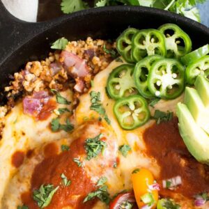 A skillet filled with a dish topped with melted cheese, sliced jalapeños, avocado, chopped cilantro, and a red sauce. There are colorful bell peppers and diced onions mixed with quinoa beneath the toppings.