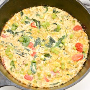 A frittata in a cast iron skillet, filled with cherry tomatoes, spinach, green onions, and melted cheese. The skillet sits on a white surface with a gray cloth partially visible in the background.