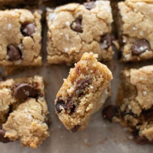 Close-up of homemade chocolate chip cookie bars, cut into squares. One piece is held upright, revealing a soft, crumbly interior with visible chocolate chips. Lightly sprinkled with sea salt flakes on top.