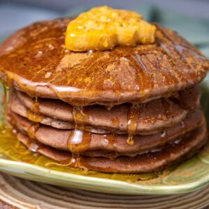A stack of pancakes on a green plate, topped with syrup and a dollop of yellow butter, sits on a wooden surface. The pancakes are richly golden, and the syrup is glistening. A green cloth is in the background.
