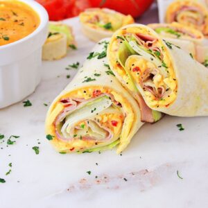 Tortilla pinwheels filled with ham, cheese, and vegetables are cut in half and served on a marble surface. A small white bowl of dipping sauce is nearby. Garnished with chopped herbs.