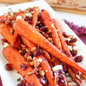 A rectangular white platter holds roasted carrots topped with crumbled cheese, pecans, dried cranberries, and fresh thyme. The dish is placed on a maroon cloth napkin, with a wooden board in the background.