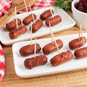 Cocktail sausages with toothpicks are arranged on two white rectangular plates. A wooden spoon and red checkered napkin are nearby. A small dish of red sauce is in the background on a wooden surface.