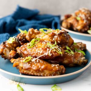 A plate of glazed chicken wings garnished with sesame seeds and sliced green onions. The wings are served on a blue plate, set on a white marble surface. A blue cloth is visible in the background.