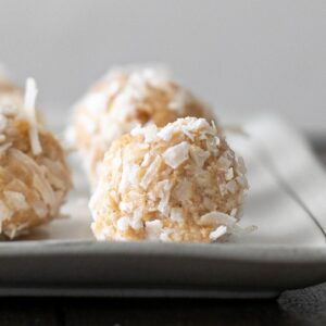 Close-up of several coconut-covered dessert balls on a white rectangular plate. They are arranged in focus in the foreground, with a blurred background, highlighting their textured appearance.