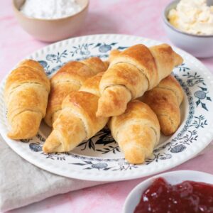 A plate of golden, freshly-baked croissants on a decorative dish. Surrounding the plate are small bowls containing butter, powdered sugar, and strawberry jam, all set on a light pink surface with a folded beige napkin nearby.