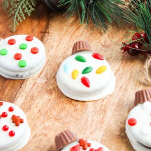 Festive cookies decorated like Christmas ornaments with white icing, red and green candies, and small candy decorations, arranged on a wooden surface with pine sprigs in the background.