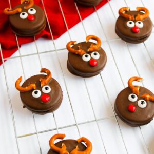 Chocolate cookies decorated as reindeer with candy eyes, red noses, and pretzel antlers are arranged on a cooling rack over a red cloth and white background.