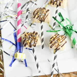 Rice Krispie treats on colorful straws, drizzled with chocolate and sprinkles, are displayed on a white plate with shiny silver and gold streamers and party blowers around them.