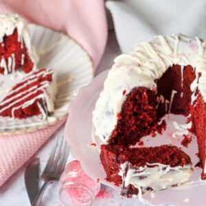A red velvet Bundt cake with white icing, partially sliced, sits on a plate. Adjacent is a separate plate with a slice of the cake, along with scattered pink sprinkles and a pink napkin in the background.