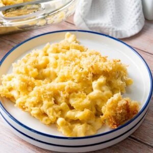 A plate of creamy macaroni and cheese with a golden breadcrumb topping sits on a wooden table. A baking dish with more macaroni and cheese and a white cloth can be seen in the background.