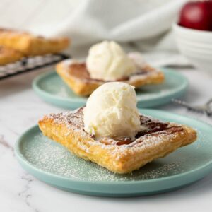 A close-up of a dessert featuring a square pastry topped with a scoop of vanilla ice cream and dusted with powdered sugar, served on a teal plate. In the background, other pastries and a bowl of red apples are visible.