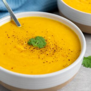 A bowl of creamy butternut squash soup, garnished with a sprig of cilantro and a sprinkle of black pepper, with a spoon in the bowl. A blue cloth is partially visible in the background.
