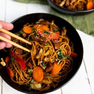A hand using chopsticks to pick up stir-fried noodles in a black bowl. The dish includes sliced carrots, bell peppers, and greens, on a white table with a green cloth visible in the background.