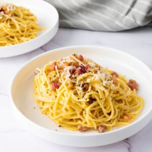 Two plates of spaghetti carbonara topped with grated cheese and bacon bits, set on a white marble surface. A striped cloth is partially visible in the background.
