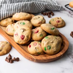 A wooden tray filled with assorted cookies containing colorful chocolate pieces, set on a marble surface. Chocolate chips are scattered around, and a gray striped cloth is draped nearby. In the background, more cookies are cooling on a wire rack.