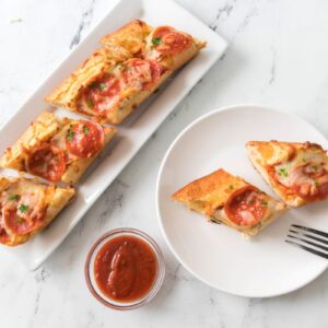 Slices of French bread pizza topped with pepperoni and cheese are arranged on a rectangular white plate. A small bowl of marinara sauce and a fork are placed next to a round white plate holding two pizza slices.