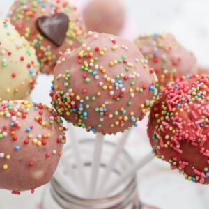 Colorful cake pops covered in pink and white coating with multicolored sprinkles, arranged in a jar. One cake pop has a small chocolate heart.