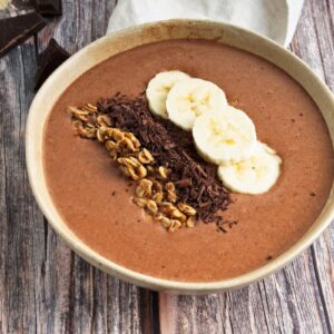 A bowl of chocolate smoothie topped with sliced bananas, granola, and shredded chocolate on a rustic wooden table.