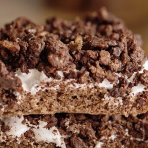 Close-up of two stacked chocolate crispy treats with a layer of sticky marshmallow. The texture appears crunchy with visible chocolate chunks and cereal pieces. The top treat has a bite taken out of it.