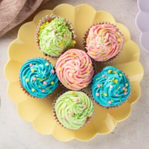 A top view of six cupcakes on a yellow scalloped plate, decorated with colorful swirl frosting in pink, blue, and green, and topped with small sprinkles. A glass bowl with sprinkles and a beige cloth are nearby on the table.