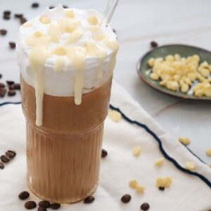 A tall glass of iced coffee topped with whipped cream and white chocolate drizzle sits on a white napkin. Coffee beans and white chocolate chips are scattered around, with additional chips in a small green dish in the background.