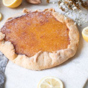 A rustic lemon tart sits on a floured surface surrounded by halved lemons and slices. Nearby are dried flowers and a rolling pin, creating a cozy baking scene. The tart has a golden-brown, crispy crust.