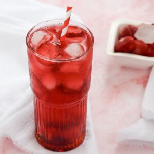 A tall glass filled with red iced drink and ice cubes, featuring a red and white striped straw, sits on a white cloth. In the background, a small dish contains frozen strawberries. The surface is a light pink marble.
