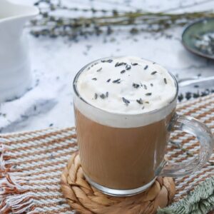 A glass mug of coffee topped with frothy cream and sprinkled with lavender sits on a woven coaster. The mug is on a striped napkin, surrounded by lavender sprigs and a small plate with more lavender. A white pitcher is in the background.