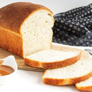 A loaf of white bread partially sliced on a wooden cutting board, accompanied by a small bowl of sauce and a serrated bread knife. A dark, patterned cloth lies beside them on a white surface.