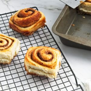 Freshly baked cinnamon rolls cooling on a wire rack, with a few remaining in a baking tray nearby. The rolls are golden brown and swirled with cinnamon, on a marble countertop. A small bowl with cinnamon is partially visible.