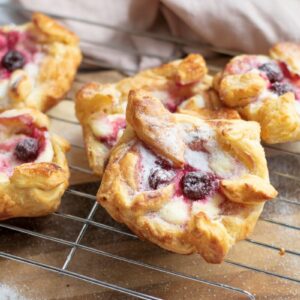 Five raspberry pastries are cooling on a wire rack. The pastries are golden brown with a flaky crust, topped with raspberries and a dusting of sugar. A light pink cloth is partially visible in the background.