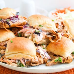 A plate of pulled pork sliders topped with coleslaw sits on an orange placemat. The buns are golden and slightly open, revealing the colorful slaw and tender pork. A wedge of lemon and a small salad are in the background.