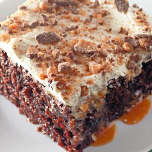 A slice of chocolate cake topped with creamy frosting and crumbled toffee pieces. Caramel drizzles over the cake and plate. The dessert sits on a white plate, highlighting the rich textures and vibrant colors.
