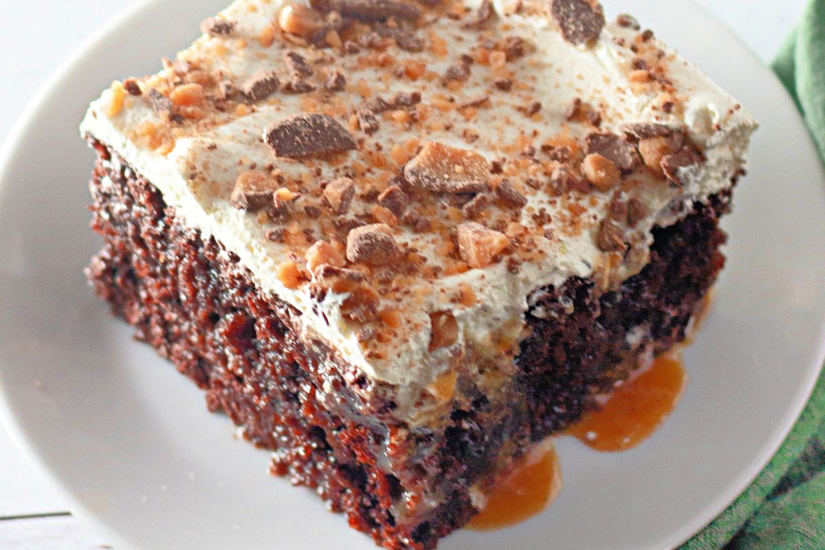 A slice of chocolate cake topped with creamy frosting and crumbled toffee pieces. Caramel drizzles over the cake and plate. The dessert sits on a white plate, highlighting the rich textures and vibrant colors.