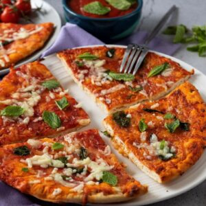 A pizza topped with cheese, basil, and spinach leaves is served on a white plate, with slices partially cut. In the background, there's a bowl of salad, a fork, cherry tomatoes, and mint leaves on a gray surface.