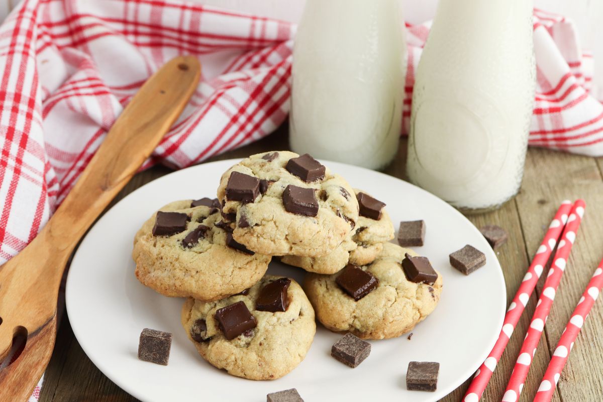 A plate of chocolate chip cookies is surrounded by chunks of chocolate. Two bottles of milk are in the background, next to red and white striped straws and a red checkered cloth. A wooden spoon rests nearby.