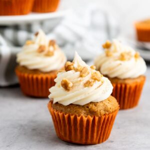 Cupcakes with white frosting and sprinkled nuts sit on a gray surface. They are in orange wrappers, and a striped cloth is partially visible in the background, along with a white cake stand holding more cupcakes.
