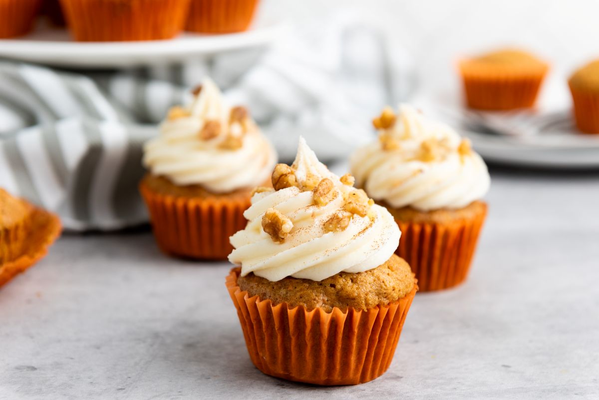 Cupcakes with white frosting and sprinkled nuts sit on a gray surface. They are in orange wrappers, and a striped cloth is partially visible in the background, along with a white cake stand holding more cupcakes.