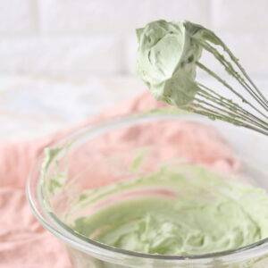 A whisk with green whipped cream is held over a glass bowl filled with the same cream. A pink cloth lies beneath the bowl on a light countertop. The background features a white tiled wall.