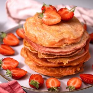 A stack of pancakes topped with sliced strawberries on a plate, surrounded by more strawberry slices. A pink napkin and a fork are visible in the background.