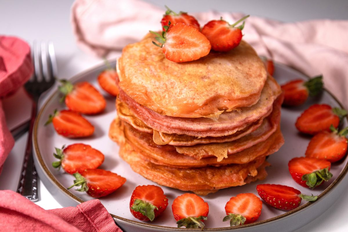 A stack of pancakes topped with sliced strawberries on a plate, surrounded by more strawberry slices. A pink napkin and a fork are visible in the background.