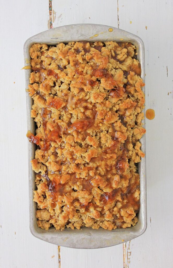 A rectangular baking pan filled with a crumbly, golden-brown streusel topping drizzled with caramel sauce, set on a white wooden surface.