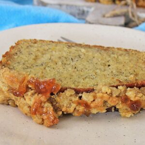 A slice of banana bread on a plate, topped with a chunky oat and raisin streusel, with a fork in the background and a blue cloth nearby.
