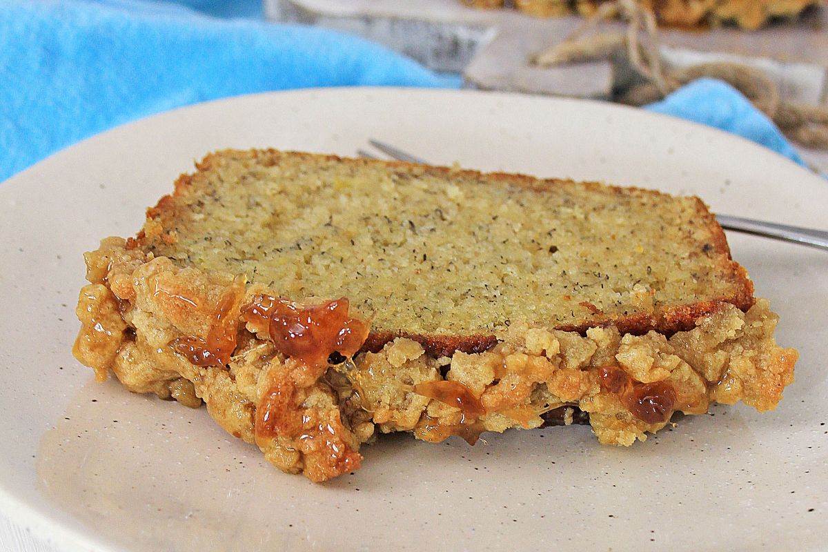 A slice of banana bread on a plate, topped with a chunky oat and raisin streusel, with a fork in the background and a blue cloth nearby.