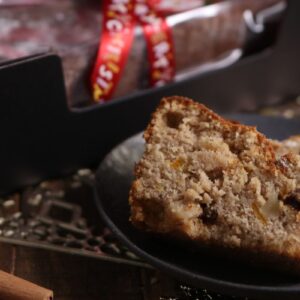 Two slices of fruit and nut cake on a dark plate, with a decorated cake box and a red ribbon in the background. Cinnamon sticks are placed nearby on a wooden surface.
