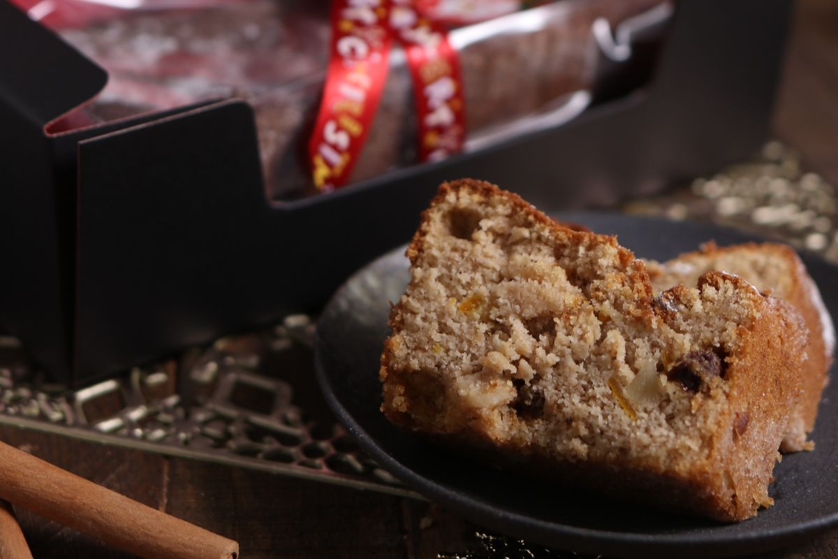 Two slices of fruit and nut cake on a dark plate, with a decorated cake box and a red ribbon in the background. Cinnamon sticks are placed nearby on a wooden surface.