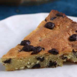 A slice of chocolate chip cookie pie on a white plate. The pie is golden brown with visible chocolate chips throughout, sitting on a deep blue surface.