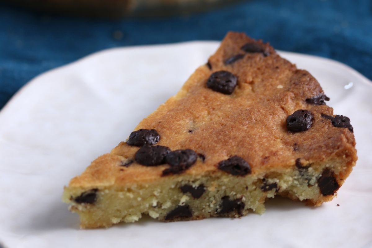 A slice of chocolate chip cookie pie on a white plate. The pie is golden brown with visible chocolate chips throughout, sitting on a deep blue surface.